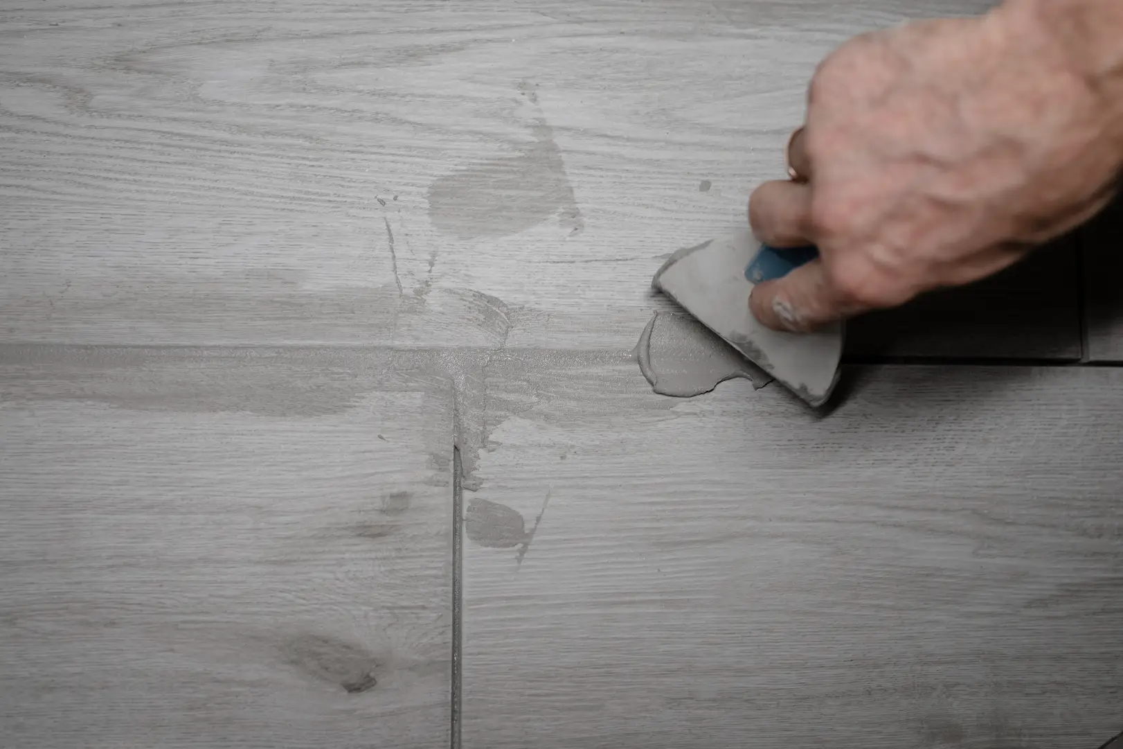 Hand uses a small tool to spread gray material along the joints of wood-look floor tiles.