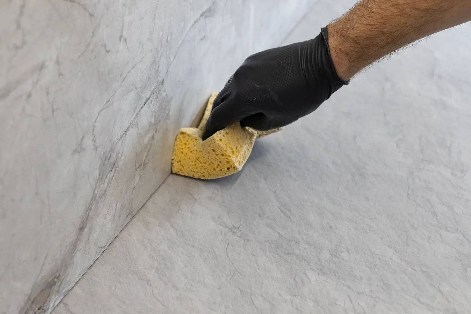 Gloved hand wipes the corner of a light gray stone-tiled surface with a yellow sponge.