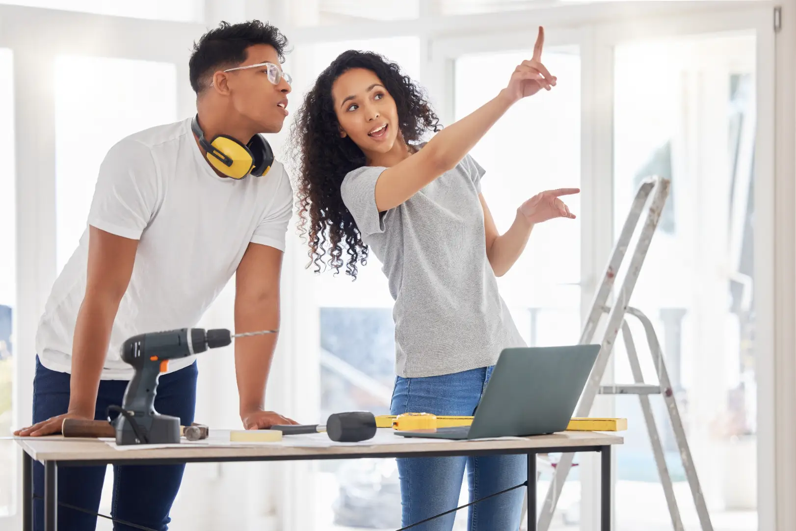 Couple stands with renovation tools, a laptop, and a ladder while pointing toward part of a bright room.