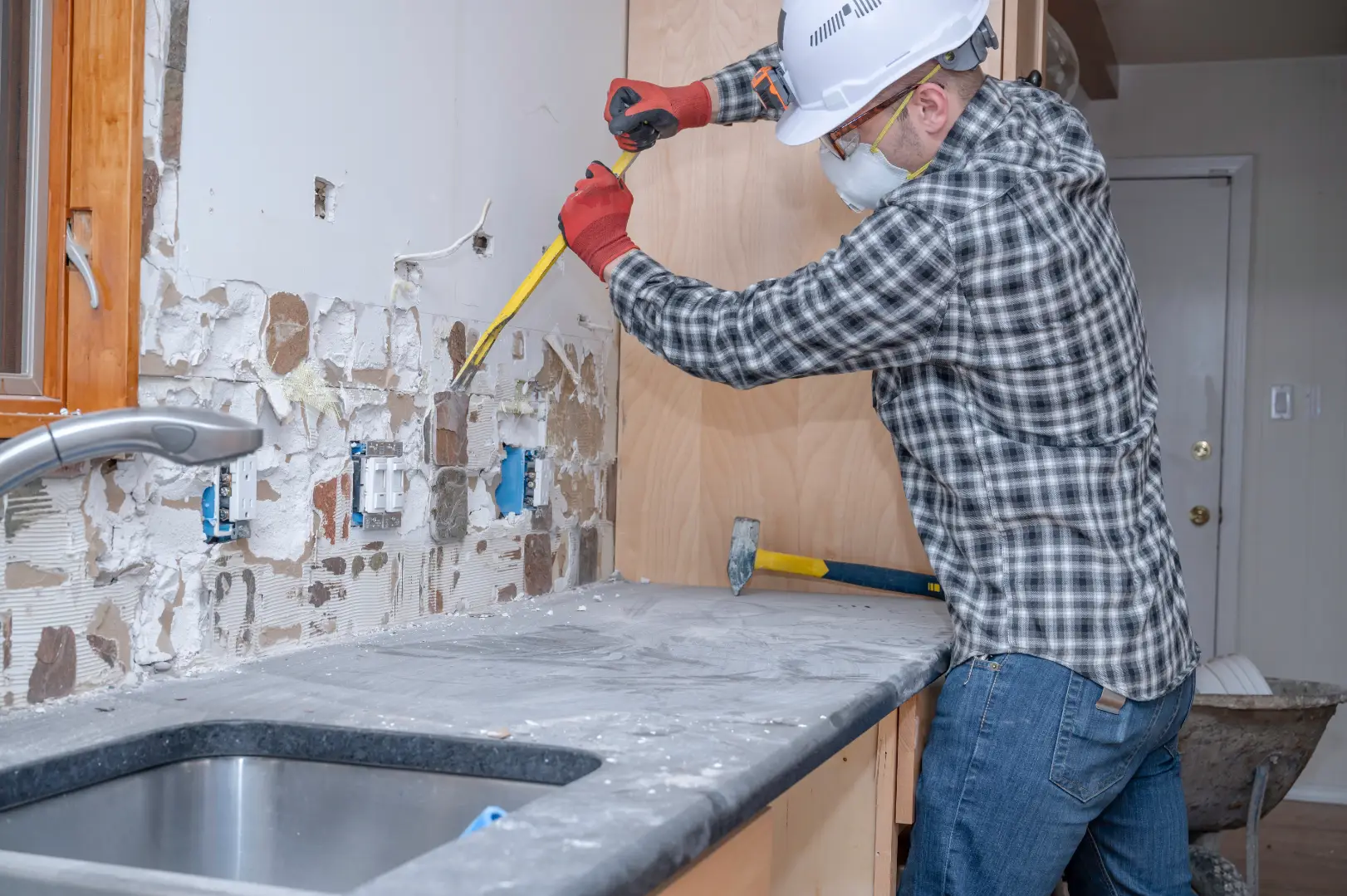 Worker in protective gear removes wall material above a kitchen countertop during renovation.